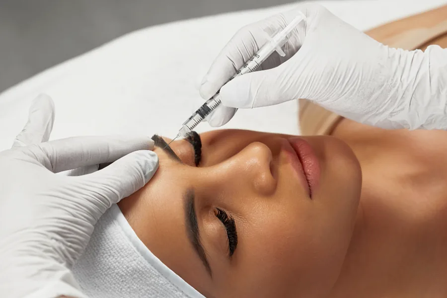 Imagem ilustrativa: profissional de luvas aplica toxina botulínica na região entre as sobrancelhas de uma mulher deitada